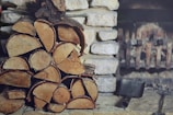 A stack of chopped firewood is neatly arranged in front of a stone wall. An old metal fireplace can be seen in the background alongside an ash shovel resting on the stone floor.