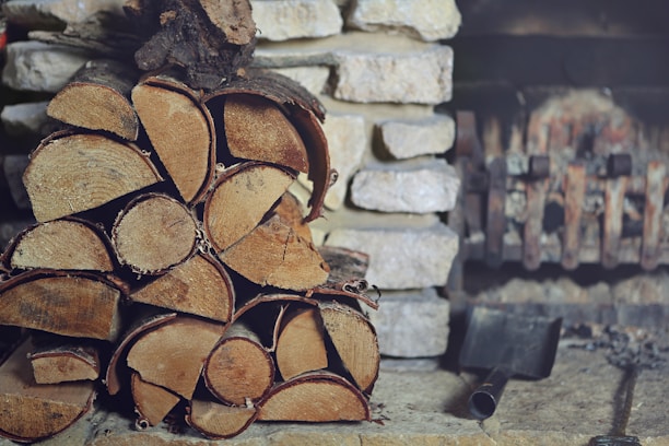 A stack of chopped firewood is neatly arranged in front of a stone wall. An old metal fireplace can be seen in the background alongside an ash shovel resting on the stone floor.