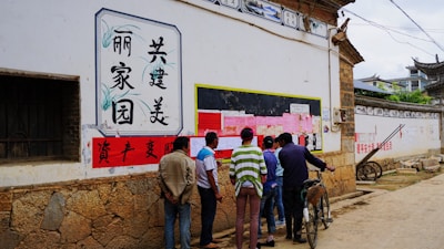 A group of people is gathered in an alleyway in front of a bulletin board covered with various pink and white papers. The wall also features Chinese characters in decorative frames. The ground is unpaved, and there's a bicycle leaning against the wall. The architecture has traditional Chinese elements, including stone and wooden structures.