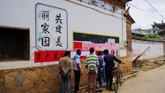A group of people is gathered in an alleyway in front of a bulletin board covered with various pink and white papers. The wall also features Chinese characters in decorative frames. The ground is unpaved, and there's a bicycle leaning against the wall. The architecture has traditional Chinese elements, including stone and wooden structures.