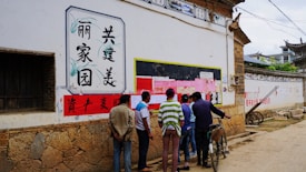 A group of people is gathered in an alleyway in front of a bulletin board covered with various pink and white papers. The wall also features Chinese characters in decorative frames. The ground is unpaved, and there's a bicycle leaning against the wall. The architecture has traditional Chinese elements, including stone and wooden structures.