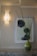 A textured wall with a geometric-patterned wall sconce emitting a warm light. Below the sconce is an abstract framed artwork featuring dark, possibly floral shapes on a light background. A glossy green vase with tall, thin grass-like plant stems is placed on a granite countertop.