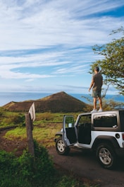 man standing on roof of Jeep Wrangler parked on dirt road