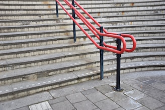 A series of concrete steps with a bright red handrail in the middle and gray brickwork in the background. The steps have tactile paving at the edges, and the ground level is paved with large concrete tiles.