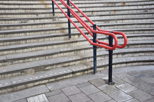 A series of concrete steps with a bright red handrail in the middle and gray brickwork in the background. The steps have tactile paving at the edges, and the ground level is paved with large concrete tiles.
