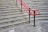 A series of concrete steps with a bright red handrail in the middle and gray brickwork in the background. The steps have tactile paving at the edges, and the ground level is paved with large concrete tiles.