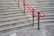 A series of concrete steps with a bright red handrail in the middle and gray brickwork in the background. The steps have tactile paving at the edges, and the ground level is paved with large concrete tiles.