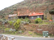 A rustic house built into a grassy hill, featuring a red roof and surrounded by stone walls and lush green vegetation. A road runs in the foreground with a waste bin next to it.