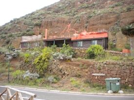 A rustic house built into a grassy hill, featuring a red roof and surrounded by stone walls and lush green vegetation. A road runs in the foreground with a waste bin next to it.