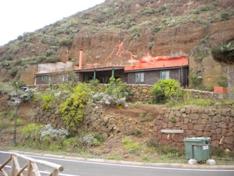 A rustic house built into a grassy hill, featuring a red roof and surrounded by stone walls and lush green vegetation. A road runs in the foreground with a waste bin next to it.