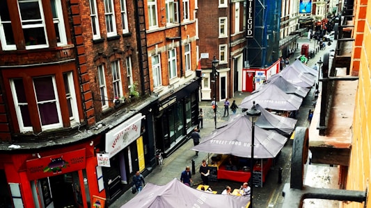 A busy urban street scene features several red brick buildings, with storefronts including a spa and a vegan restaurant. Along the street, a row of gray canopies cover street food stalls. People are walking along the pavement, and the area has a vibrant, bustling atmosphere.