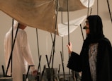 Two individuals in traditional attire are interacting with an art installation composed of vertical poles and fabric. The setting suggests an indoor environment, possibly an art gallery.