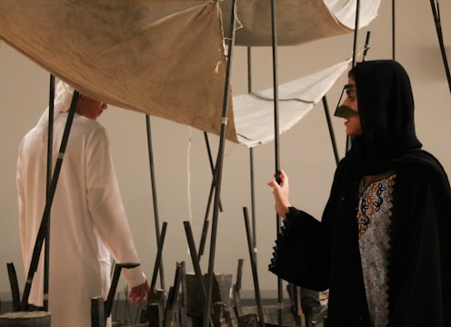 Two individuals in traditional attire are interacting with an art installation composed of vertical poles and fabric. The setting suggests an indoor environment, possibly an art gallery.