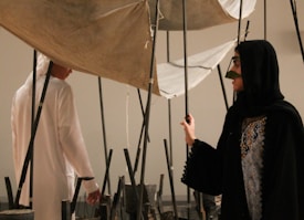 Two individuals in traditional attire are interacting with an art installation composed of vertical poles and fabric. The setting suggests an indoor environment, possibly an art gallery.