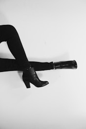 Two crossed legs wearing black shoes against a plain white background. The image captures a modern, minimalist fashion style with high-heeled ankle boots and slim-fitting pants.