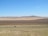 A landscape featuring vast, open plains with different shades of brown and green fields. In the distance, there are rolling hills or low mountains under a clear blue sky. In the foreground, an isolated vehicle travels along a road.