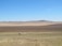 A landscape featuring vast, open plains with different shades of brown and green fields. In the distance, there are rolling hills or low mountains under a clear blue sky. In the foreground, an isolated vehicle travels along a road.