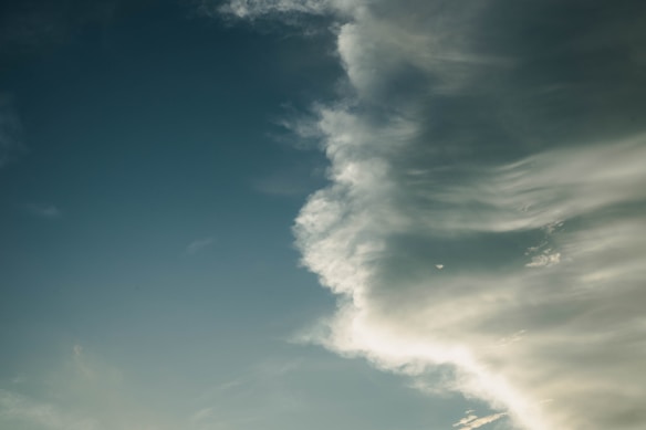 A sky scene with a dramatic formation of white clouds sweeping across from the right side against a backdrop of deep blue sky. The clouds appear soft and wispy, illuminated by sunlight.