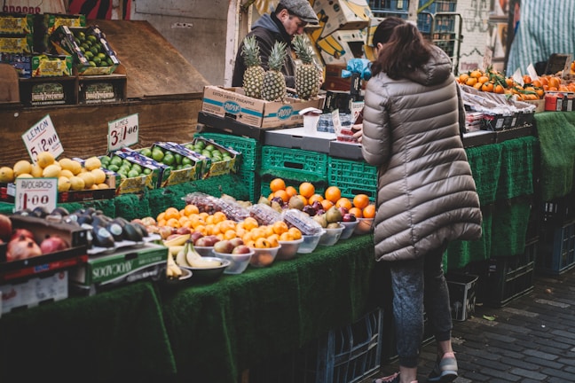 A bustling market stall displays a variety of fresh fruits. There are limes, avocados, oranges, apples, and pineapples neatly arranged on the table. A woman wearing a beige puffer jacket is examining the produce. Behind the stall, a vendor in a cap is attending to the goods. The market area is covered with green cloth, enhancing the vibrant colors of the fruits.