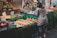 A bustling market stall displays a variety of fresh fruits. There are limes, avocados, oranges, apples, and pineapples neatly arranged on the table. A woman wearing a beige puffer jacket is examining the produce. Behind the stall, a vendor in a cap is attending to the goods. The market area is covered with green cloth, enhancing the vibrant colors of the fruits.
