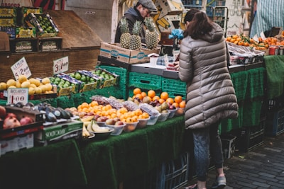 A bustling market stall displays a variety of fresh fruits. There are limes, avocados, oranges, apples, and pineapples neatly arranged on the table. A woman wearing a beige puffer jacket is examining the produce. Behind the stall, a vendor in a cap is attending to the goods. The market area is covered with green cloth, enhancing the vibrant colors of the fruits.