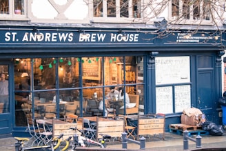 A brew house with a dark exterior and large glass windows displaying a cozy interior. Inside, there are tables and chairs with people sitting and enjoying drinks. The establishment advertises brewery tours and quiz nights on handwritten signs. Outside, a few bicycles are parked on the street.