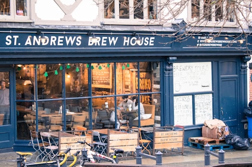 A brew house with a dark exterior and large glass windows displaying a cozy interior. Inside, there are tables and chairs with people sitting and enjoying drinks. The establishment advertises brewery tours and quiz nights on handwritten signs. Outside, a few bicycles are parked on the street.