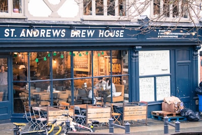 A brew house with a dark exterior and large glass windows displaying a cozy interior. Inside, there are tables and chairs with people sitting and enjoying drinks. The establishment advertises brewery tours and quiz nights on handwritten signs. Outside, a few bicycles are parked on the street.