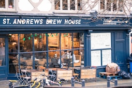 A brew house with a dark exterior and large glass windows displaying a cozy interior. Inside, there are tables and chairs with people sitting and enjoying drinks. The establishment advertises brewery tours and quiz nights on handwritten signs. Outside, a few bicycles are parked on the street.
