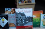 A collection of books displayed upright in a bookstore. Prominent titles include 'Rural Britain Then & Now' featuring a cover image of traditional thatched cottages, 'The Baking Bible' with images of eggs and a pie crust, and 'Home and Fashion A Concise History' with a stylized illustration of a woman in a dress. The books are neatly arranged on a shelf with price tags visible.