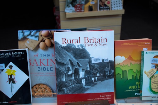 A collection of books displayed upright in a bookstore. Prominent titles include 'Rural Britain Then & Now' featuring a cover image of traditional thatched cottages, 'The Baking Bible' with images of eggs and a pie crust, and 'Home and Fashion A Concise History' with a stylized illustration of a woman in a dress. The books are neatly arranged on a shelf with price tags visible.