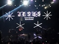 A stage with bright lights and a musical band performing. Large text displaying 'JESUS' and smaller text stating 'Yours is the Name Above all names' is visible on the screen. The crowd appears to be engaged in the performance.