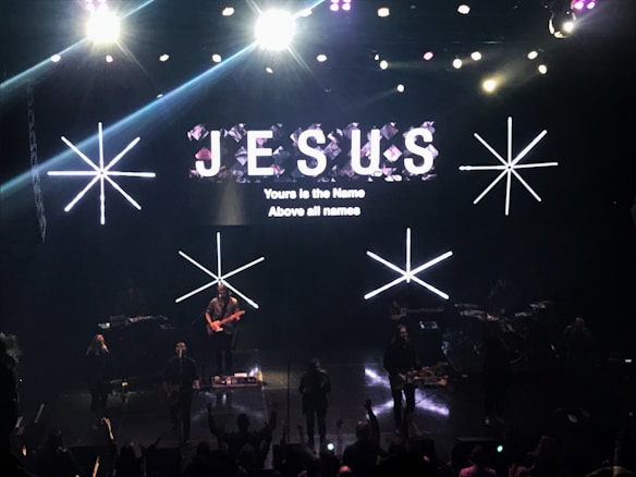 A stage with bright lights and a musical band performing. Large text displaying 'JESUS' and smaller text stating 'Yours is the Name Above all names' is visible on the screen. The crowd appears to be engaged in the performance.