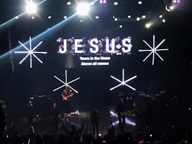 A stage with bright lights and a musical band performing. Large text displaying 'JESUS' and smaller text stating 'Yours is the Name Above all names' is visible on the screen. The crowd appears to be engaged in the performance.