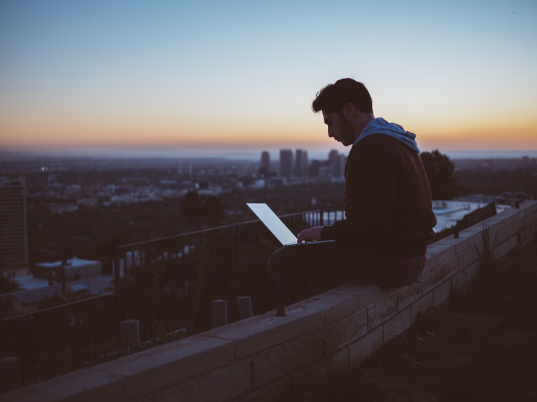 A young man working on a laptop while seated on a ledge in a city during sunset