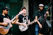 Three musicians are passionately performing outdoors. One plays an acoustic guitar, another a banjo, and the third a double bass. They are casually dressed in t-shirts and jeans, contributing an informal, street music vibe. The expressions on their faces indicate immersion in their music, with one singing along.