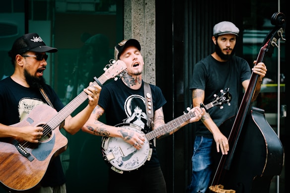 Three musicians are passionately performing outdoors. One plays an acoustic guitar, another a banjo, and the third a double bass. They are casually dressed in t-shirts and jeans, contributing an informal, street music vibe. The expressions on their faces indicate immersion in their music, with one singing along.