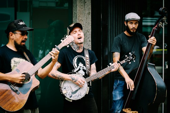 Three musicians are passionately performing outdoors. One plays an acoustic guitar, another a banjo, and the third a double bass. They are casually dressed in t-shirts and jeans, contributing an informal, street music vibe. The expressions on their faces indicate immersion in their music, with one singing along.