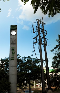 A tall concrete pillar with a clock near the top stands beside a set of utility poles. Lush green trees are in the foreground, and a blue sky with scattered clouds can be seen overhead. The word 'Itagui' is vertically inscribed on the pillar.