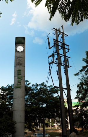 A tall concrete pillar with a clock near the top stands beside a set of utility poles. Lush green trees are in the foreground, and a blue sky with scattered clouds can be seen overhead. The word 'Itagui' is vertically inscribed on the pillar.