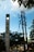 A tall concrete pillar with a clock near the top stands beside a set of utility poles. Lush green trees are in the foreground, and a blue sky with scattered clouds can be seen overhead. The word 'Itagui' is vertically inscribed on the pillar.