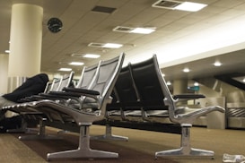 Rows of empty airport seats with a person lying across them on the left, wearing dark clothing. A clock is visible on a pillar, and the ceiling features recessed lighting panels. The floors are carpeted, and the overall environment appears quiet and subdued.