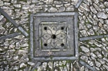 A detailed paving pattern with small, irregular stones arranged around a decorative, square-shaped drain cover. The drain cover features a floral design with symmetrical petal-like shapes and circular openings. The stones are interspersed with patches of moss, adding texture and natural elements to the scene.