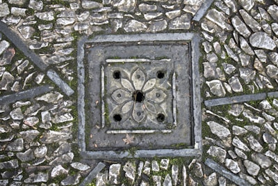 A detailed paving pattern with small, irregular stones arranged around a decorative, square-shaped drain cover. The drain cover features a floral design with symmetrical petal-like shapes and circular openings. The stones are interspersed with patches of moss, adding texture and natural elements to the scene.