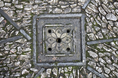 A detailed paving pattern with small, irregular stones arranged around a decorative, square-shaped drain cover. The drain cover features a floral design with symmetrical petal-like shapes and circular openings. The stones are interspersed with patches of moss, adding texture and natural elements to the scene.