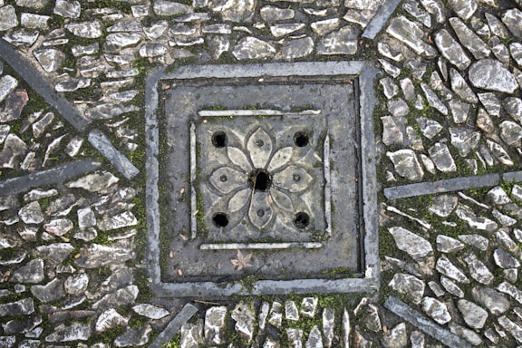A detailed paving pattern with small, irregular stones arranged around a decorative, square-shaped drain cover. The drain cover features a floral design with symmetrical petal-like shapes and circular openings. The stones are interspersed with patches of moss, adding texture and natural elements to the scene.