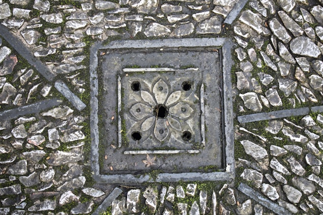 A detailed paving pattern with small, irregular stones arranged around a decorative, square-shaped drain cover. The drain cover features a floral design with symmetrical petal-like shapes and circular openings. The stones are interspersed with patches of moss, adding texture and natural elements to the scene.