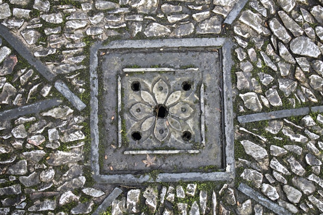 A detailed paving pattern with small, irregular stones arranged around a decorative, square-shaped drain cover. The drain cover features a floral design with symmetrical petal-like shapes and circular openings. The stones are interspersed with patches of moss, adding texture and natural elements to the scene.