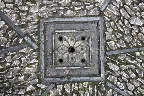 A detailed paving pattern with small, irregular stones arranged around a decorative, square-shaped drain cover. The drain cover features a floral design with symmetrical petal-like shapes and circular openings. The stones are interspersed with patches of moss, adding texture and natural elements to the scene.
