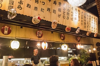 A cozy restaurant interior with a warm, ambient lighting and a wooden panel adorned with masks and Japanese calligraphy. Lanterns hang from the ceiling, and a chef is visible behind a counter, interacting with customers.